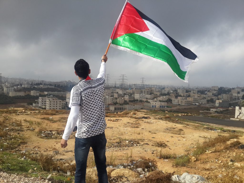 A man waving a Palestinian flag standing on a hill overlooking a city in Gaza.