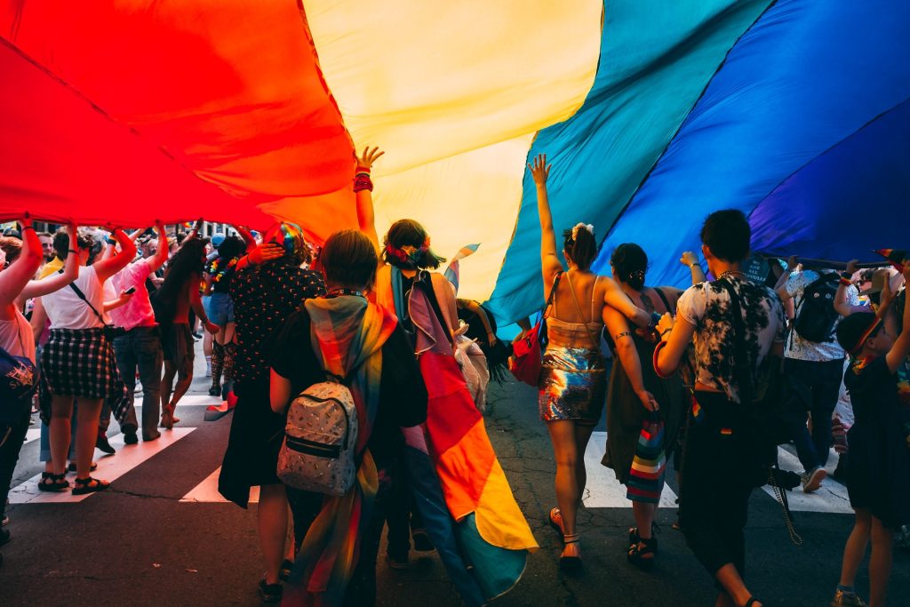 A pride march with people walking under a giant parachute style rainbow banner.