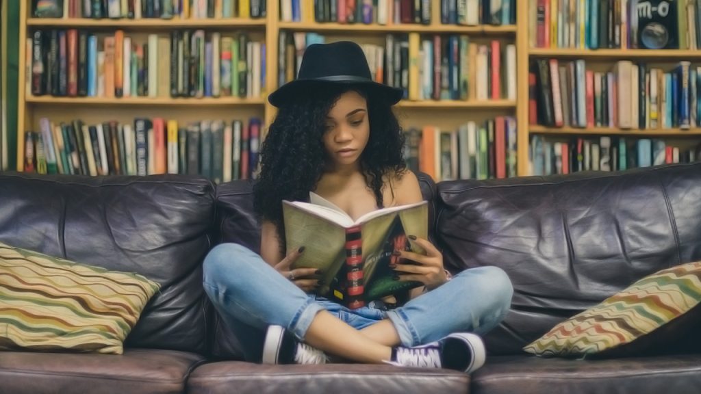 A trendy girl sitting crosslegged on a leather sofa reading a book with shelves of books in the background.