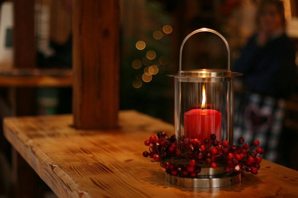 A candle in a lantern surrounded by red berries sitting on a wooden shelf in a warm glowing room at night.