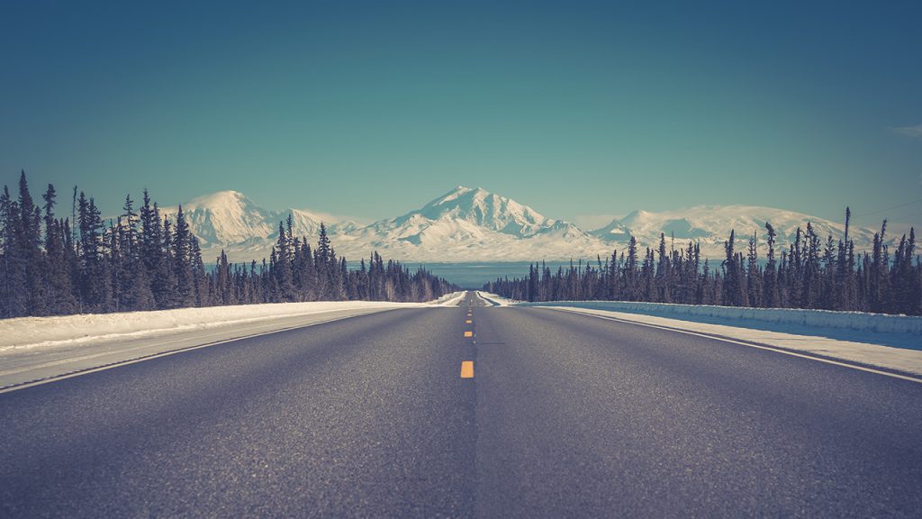 A long, wide, straight road going off into the distance with a pine forest either side and snow-capped mountains in the distance.