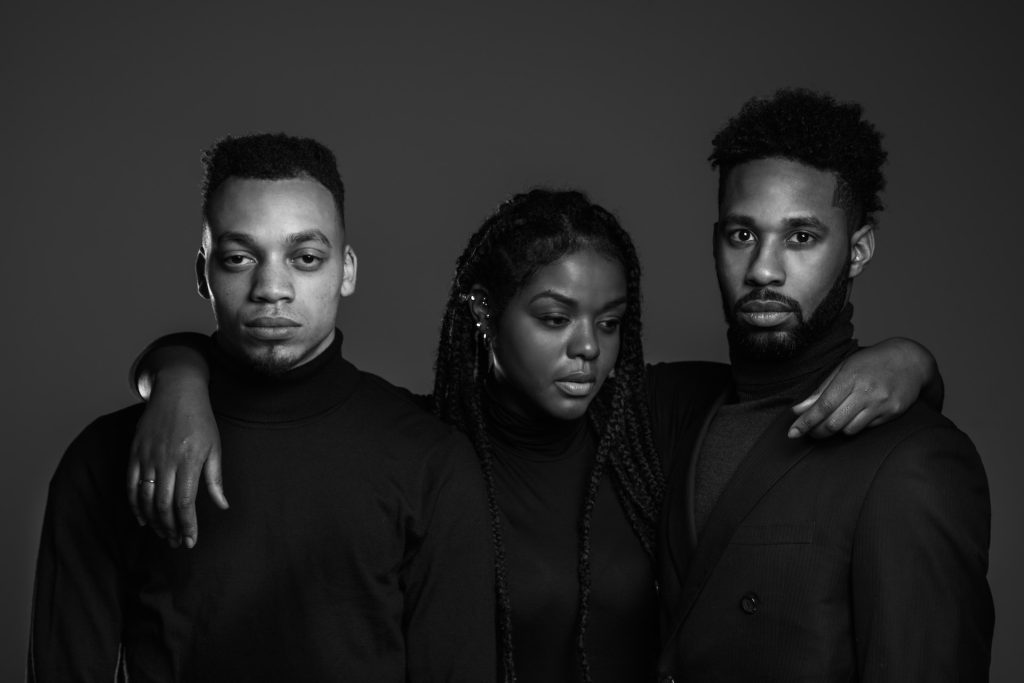 Three stunning and stylish black people - a woman in the centre with her hands draped around the shoulders of the two men.