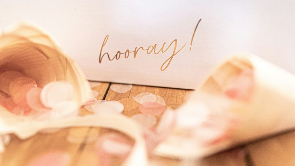 A card standing next to confetti with the word 'hooray' written on it in gold writing.