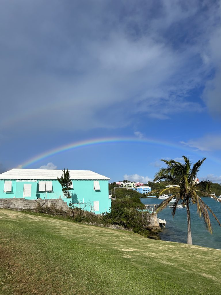 A rainbow over a waterside Bermuda setting with a pastel-coloured house and a palm tree.