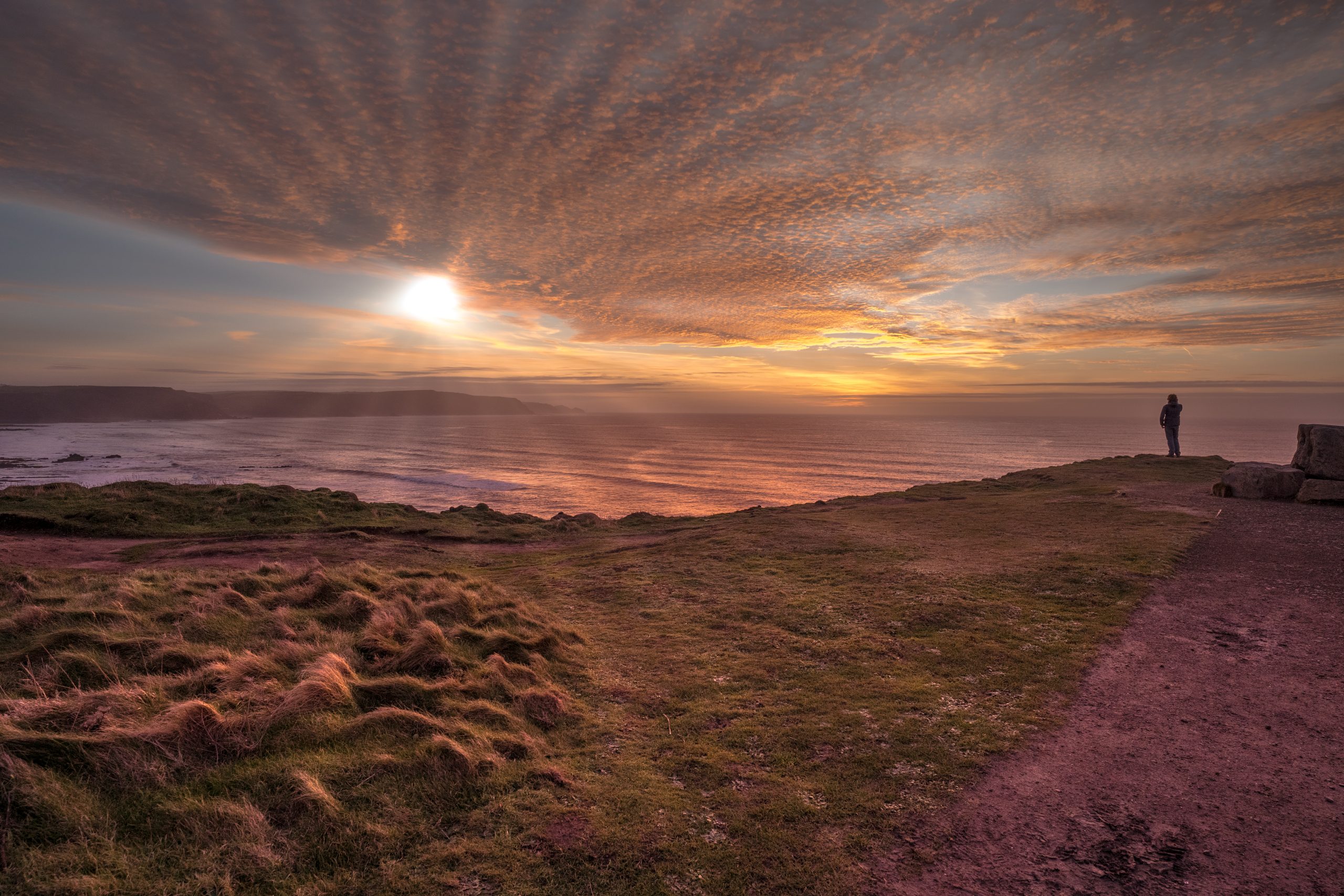 A person standing on a cliff at sunset looking at the sea and sky.