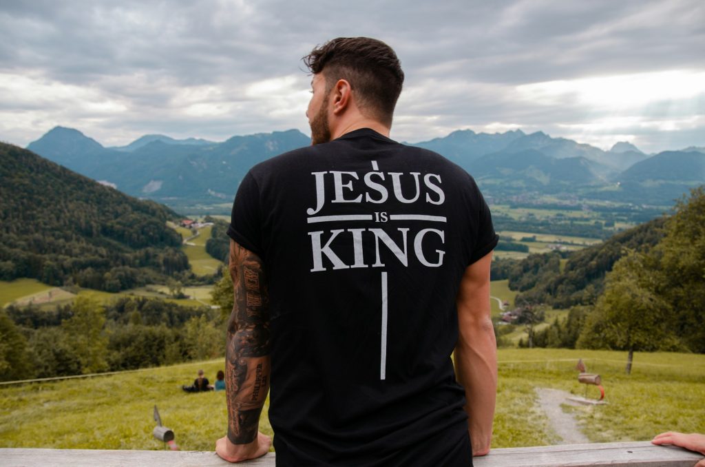 A tattooed man looking out of a vista with a t-shirt with Jesus is King written on the back.