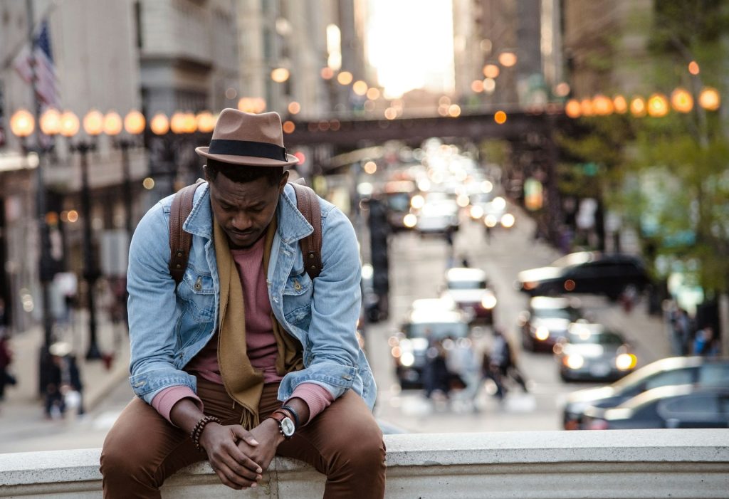 A man in a hat with a backpack sitting on a bridge looking sown and sad with an urban evening street scene behind him.