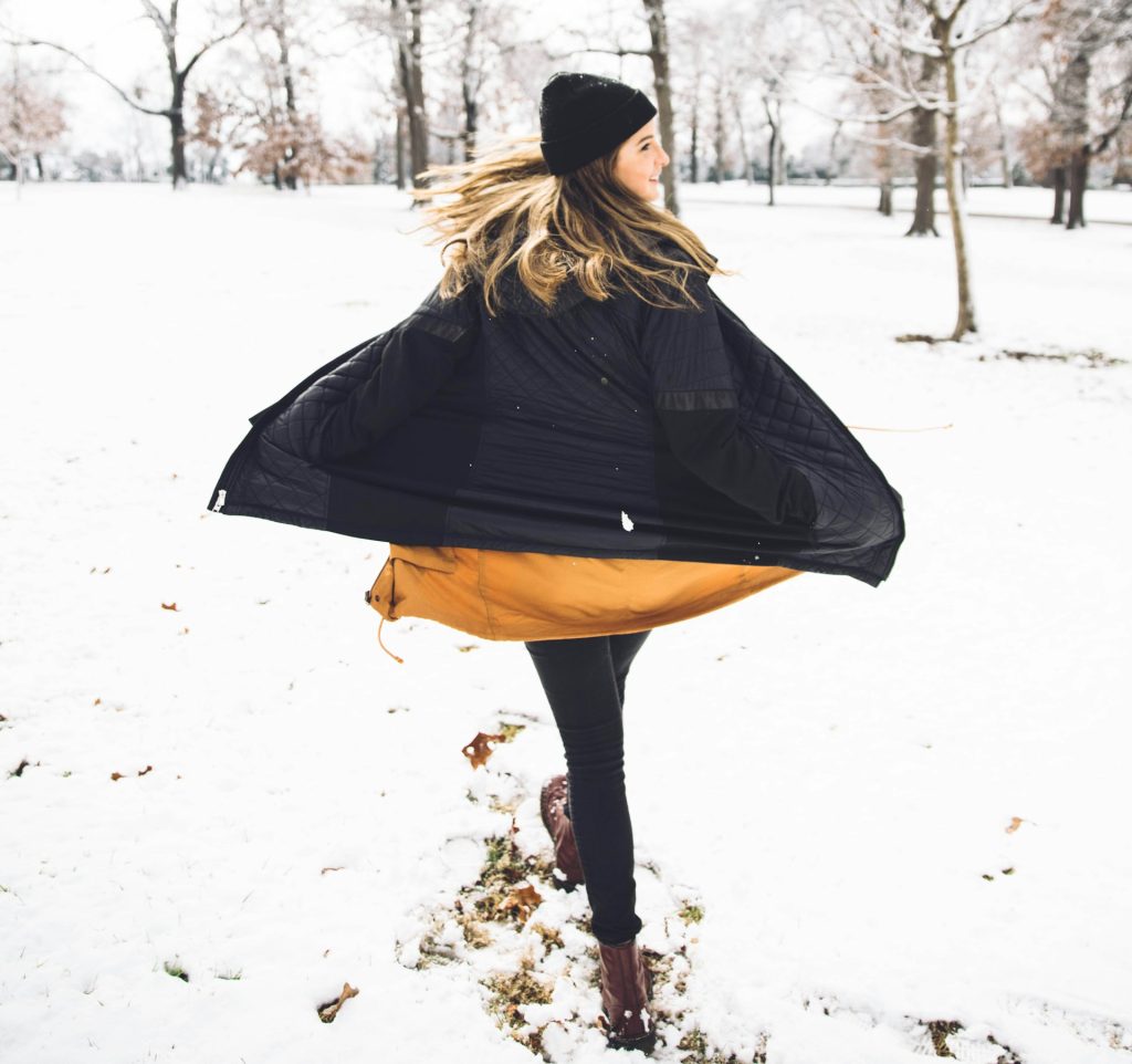 A woman with a hat and her hands in her pockets swirling her coat in a snowy forest.