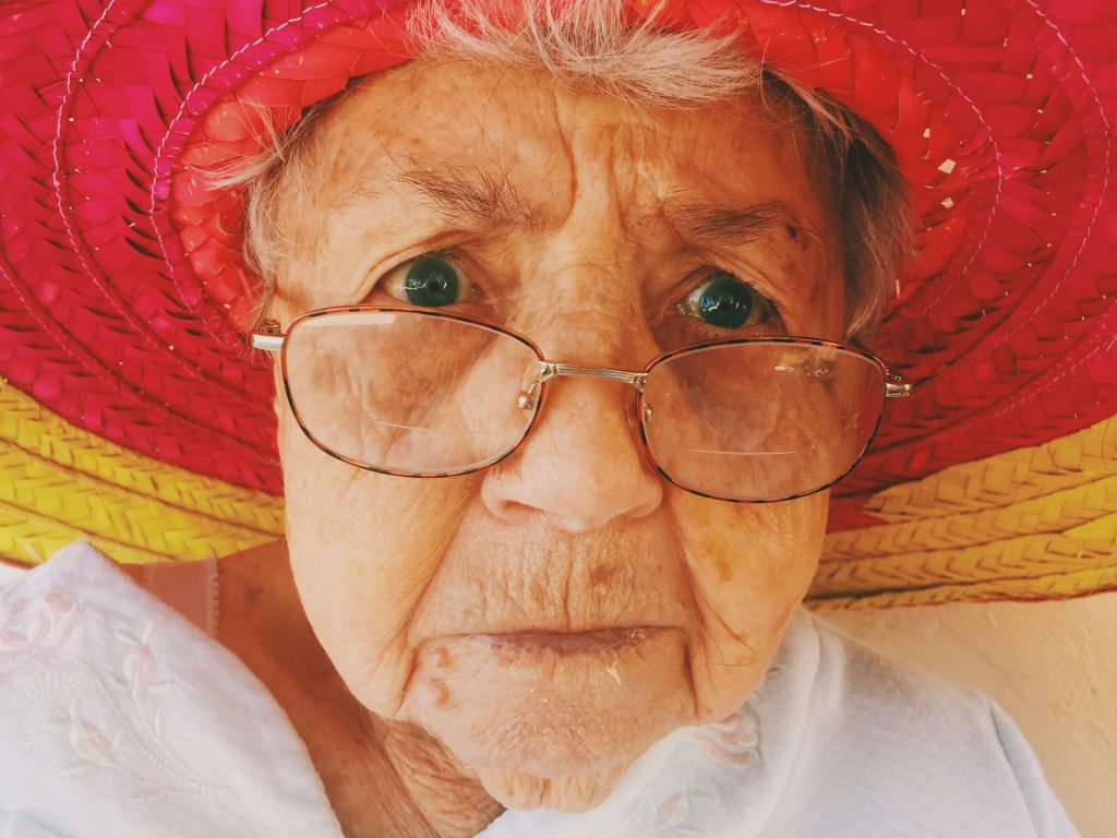 The close-up of a grumpy looking old woman looking over the top of her glasses and wearing a wide-brimmed hat.