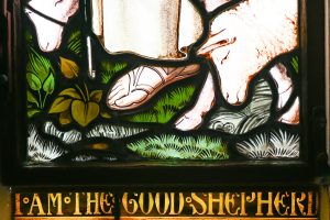 A closeup of a stained-glass window with the feet of Jesus and a sheep with the words underneath, I am the good shepherd.