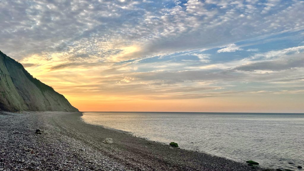 A beach with cliff and a clouded sky with the sun rising out of sight behind the cliff.