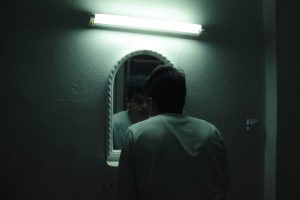A man in a dark room leaning in to see his reflection in a mirror.