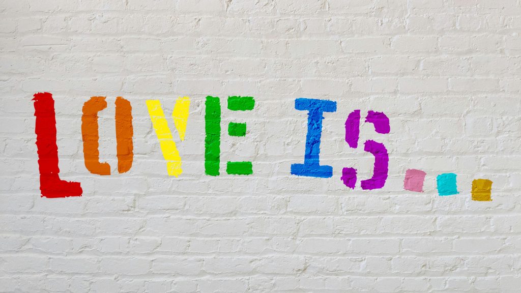 A blank brick wall with the words 'Love is' stencilled with each of the letters in a different colour of the Pride flag.