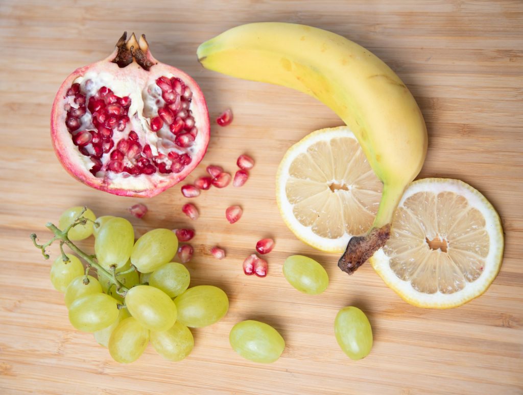 A mixture of fruits whole and halved showing their interiors. pomegranate, banana, lemon, and grapes.