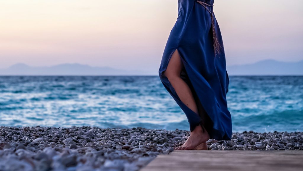 A person stands on a pebbly beach by the sea at sunset and is wearing a long blue dress.