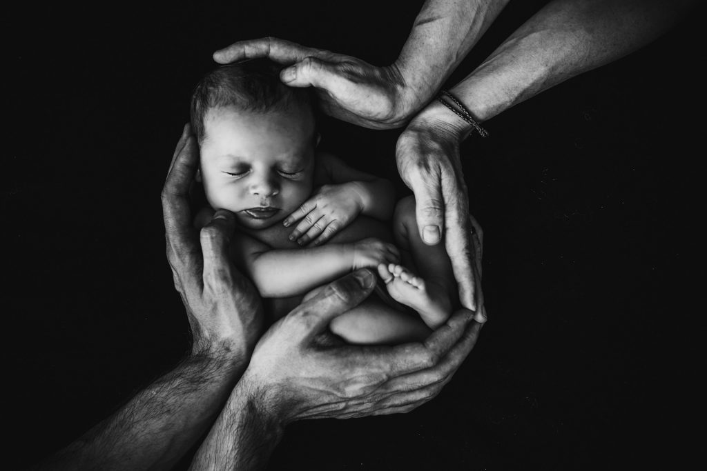 A male and female pair of hands form a cradle around a baby.