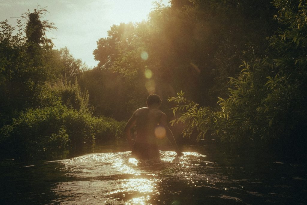 A man stands waist deep in a small river with his back to the camera. The banks are covered in lush and thick green undergrowth and trees.