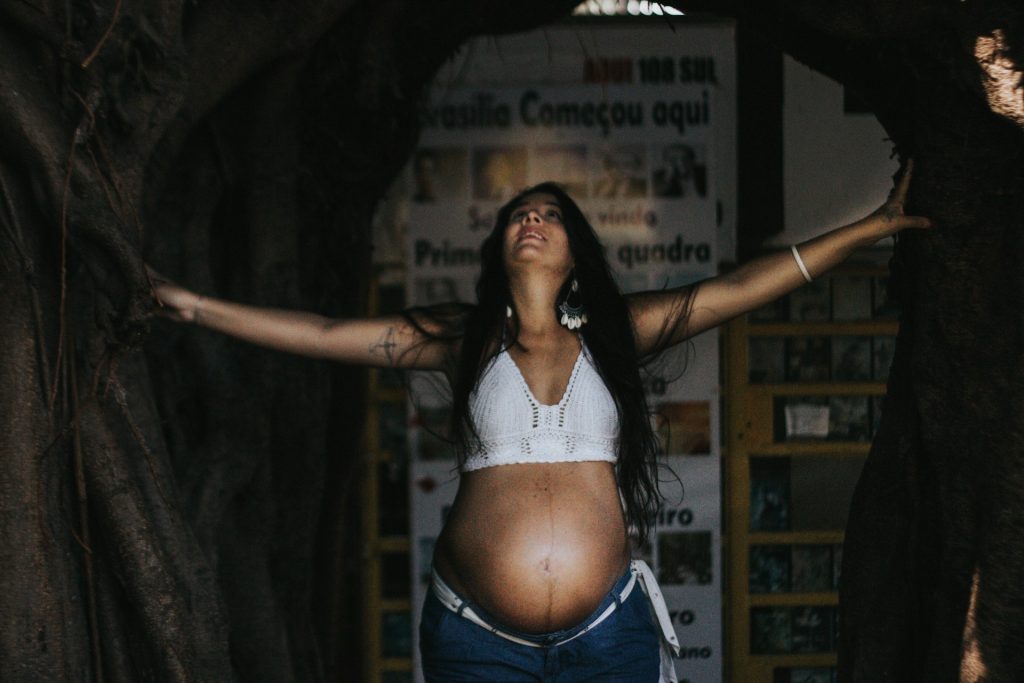 A heavily pregnant woman stands in room holding back two curtains with her hands, her belly exposed, whilst smiling and looking up with joy.