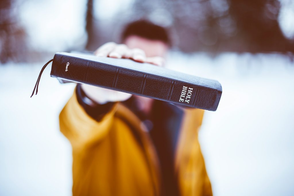 A man, standing in the snow, holds a Bible towards the camera.