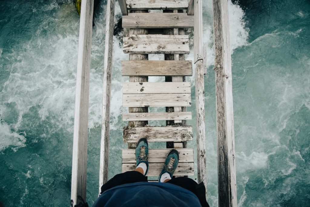 Looking down on a rickety old bridge spanning a raging river, a person is stepping out onto the wooden planks.