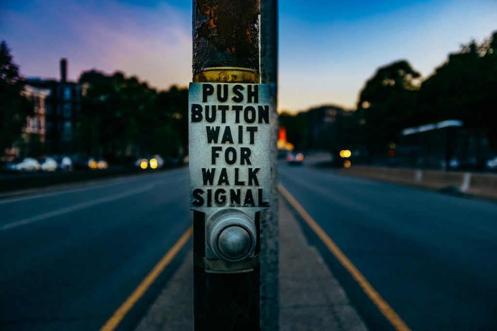 An evening at sunset. A post in the central island between road highways - on a bysy street - has a rusted metal sign and a button. The sign reads, "PUSH BUTTON WAIT FOR WALK SIGNAL".