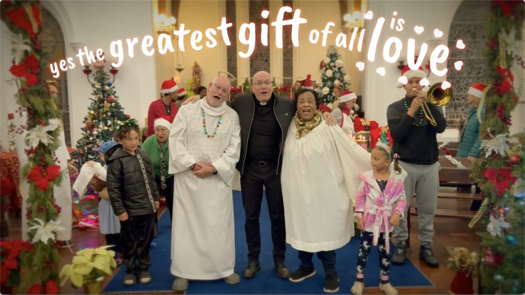 A screenshot of the video showing the whole cast of the Christmas video with the words The Greatest Gift of All is Love