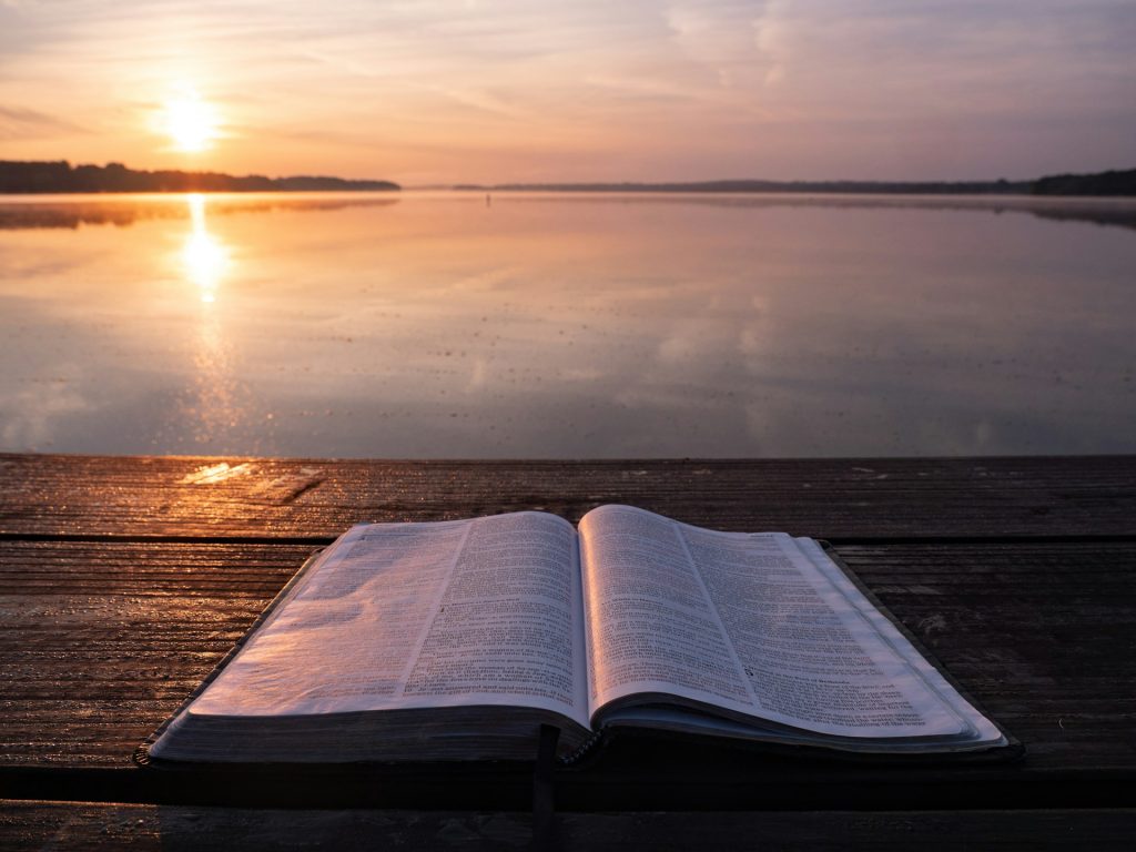 A Bible lays open on a wooden dock as the sun rises over a lake.