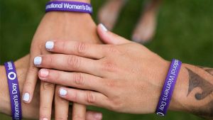 Three women's hands being held out and placed on top of each other, each with a rubber purple International Womens' Day bracelet.