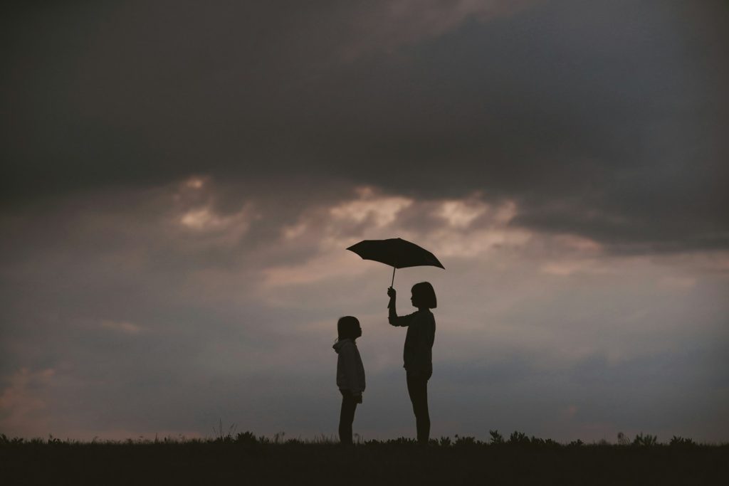 Two people stand in a field silhouetted against a cloudy sky with a person holding an umbrella over a child.