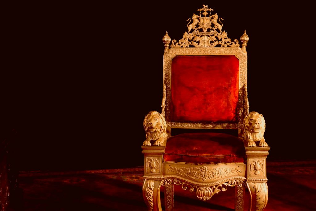 A beautiful and elaborate golden throne with red velvet seat and back sits on a plinth in a darkened room.
