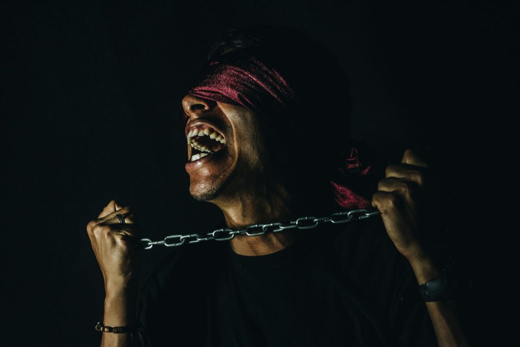 A man stands in the darkness, blindfolded and holding between his hands a chain. He is shouting out in anguish.