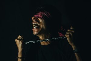 A man stands in the darkness, blindfolded and holding between his hands a chain. He is shouting out in anguish.
