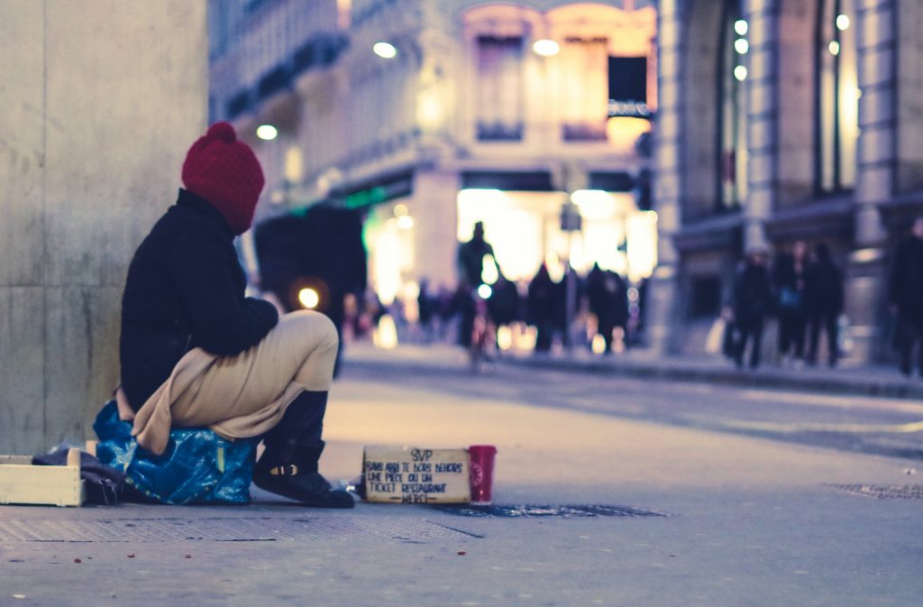 A homeless person sits alone on a street corner at night, illuminated by the glow from restaurants and shops. Used under license from EV on Unsplash.