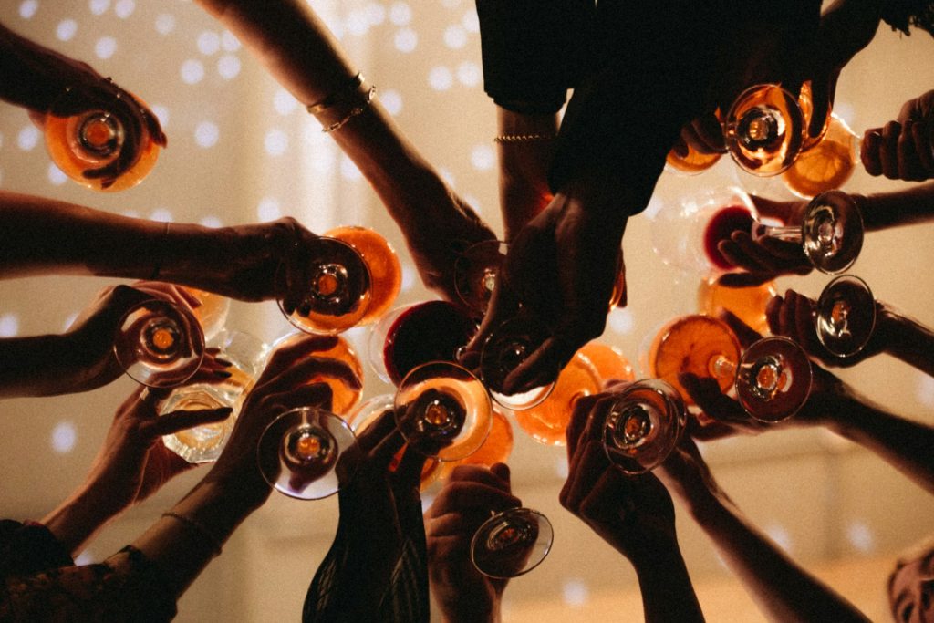 A group of people chink their drinks together and the view is underneath looking upwards at all their arms being stretched out and their hands holding the glasses.