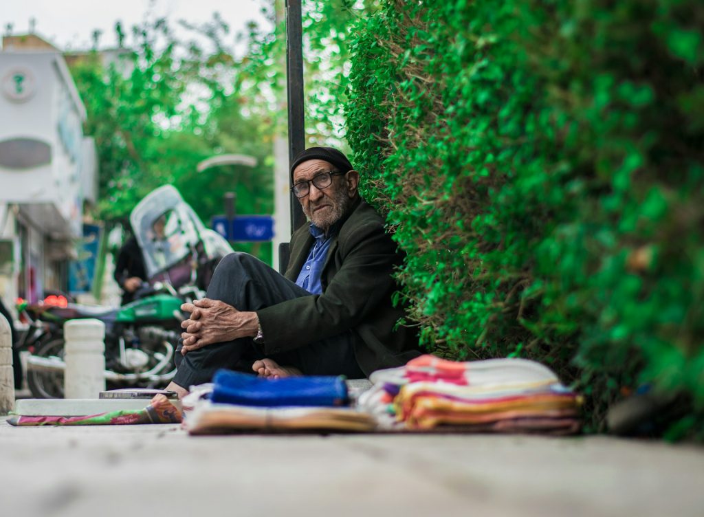A homeless man, sitting on the ground, looks directly into the camera.