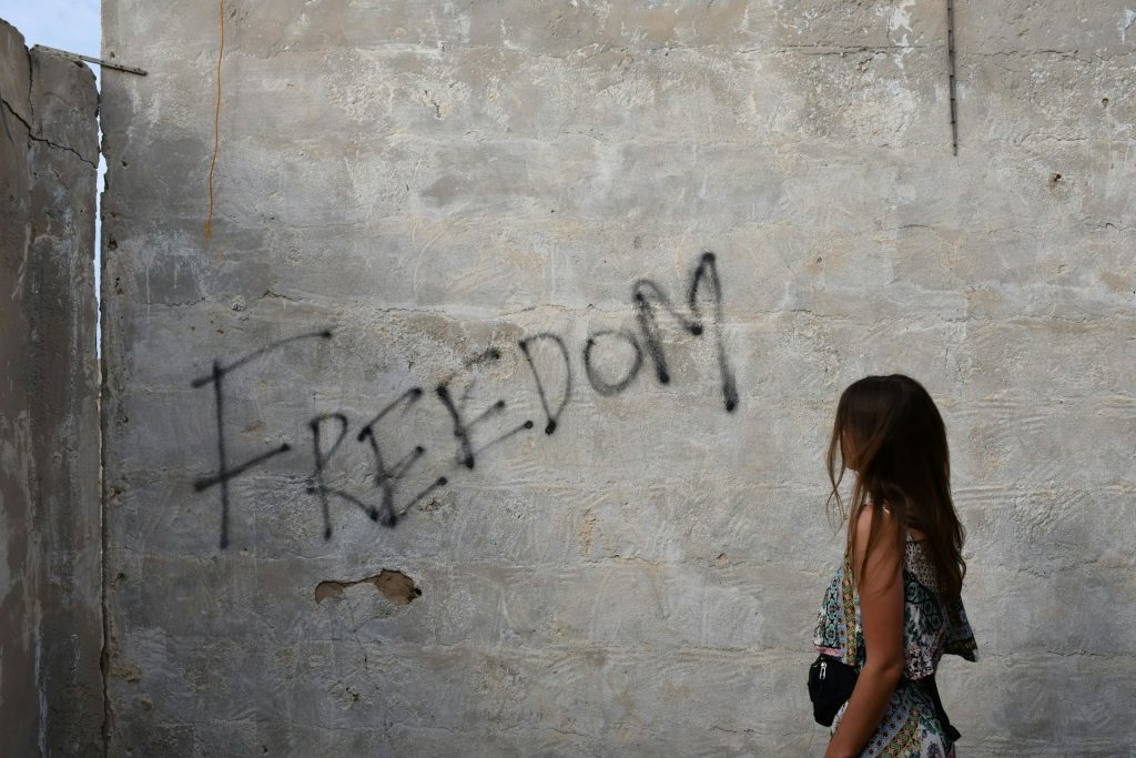 A woman stands, staring at a wall with the word 'FREEDOM' spray-painted onto it.