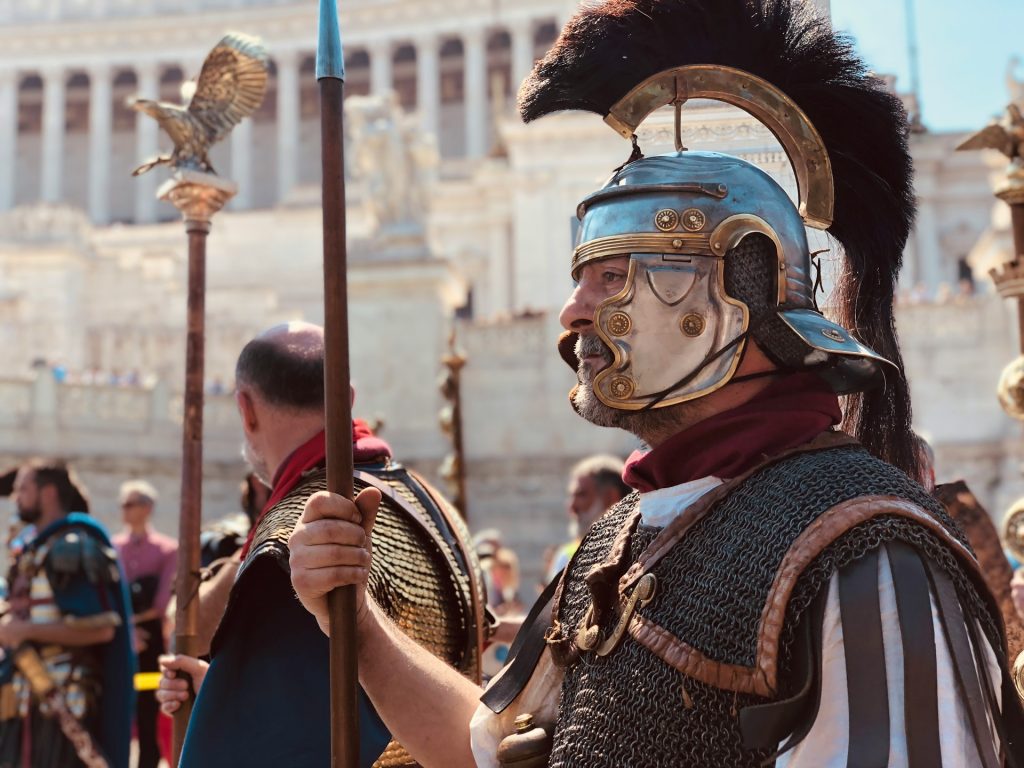 Roman soldiers stand to attention with their full armour, helmets, and staffs.