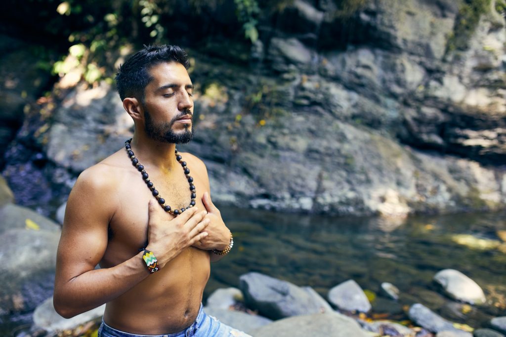 A man, bare-chested, stands by a forest river with his hands on his heart and his eyes closed in meditative prayer.