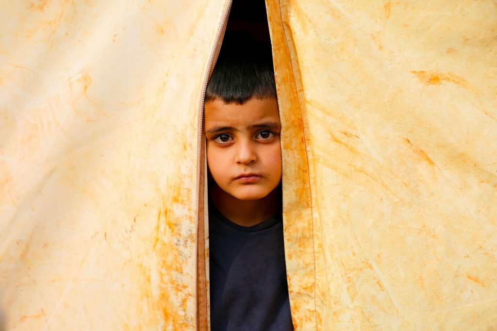 A young, sad-looking boy, pokes his head through a gap in a yellowing, dirty, canvas tent.