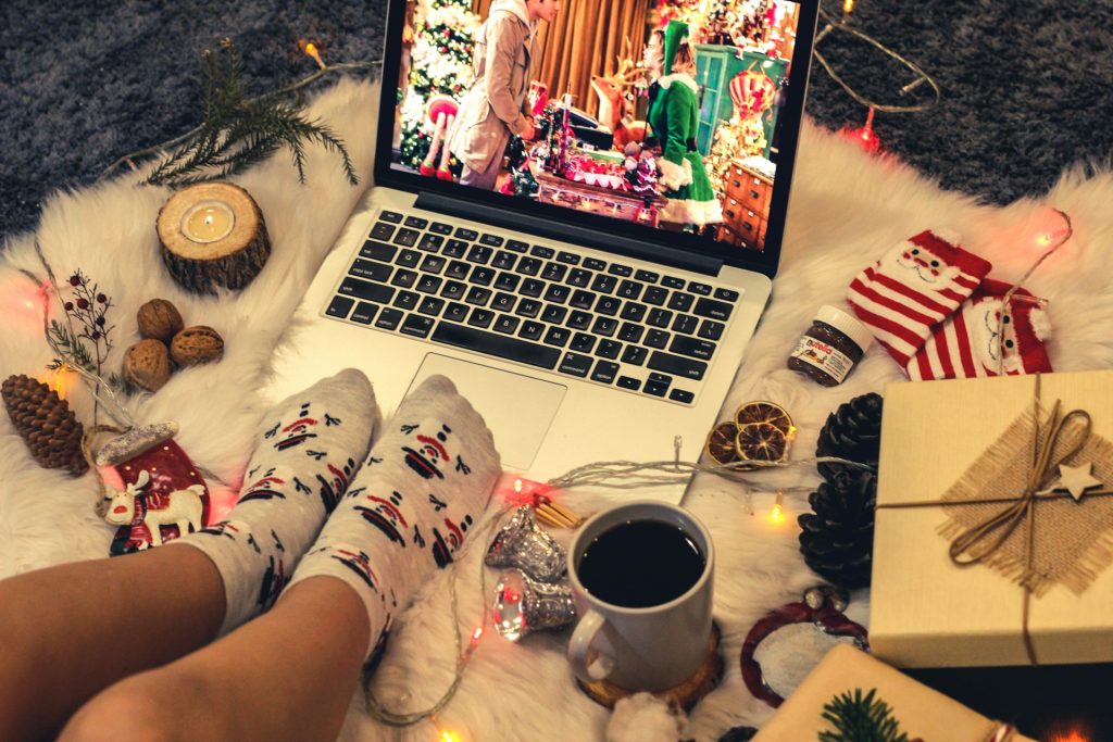 A person wearing Christmas socks watches a Christmas movie on their lapop. The floor around them is littered with Chrismassy things.