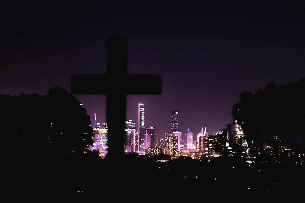 A cross in the foreground is silhouetted against an illuminated night-time cityscape.