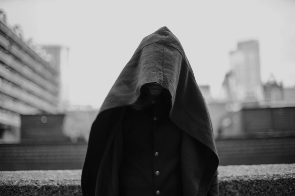 A hooded figure (a person of unknown gender) stands on a rooftop alone before a cityscape.