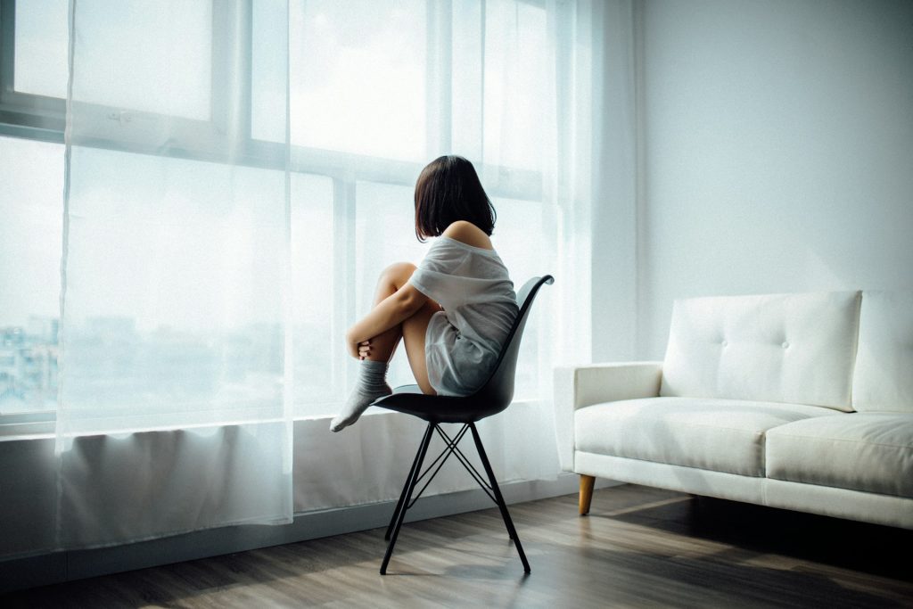 A woman sits alone on a chair, hugging herself, and looking out of a window.