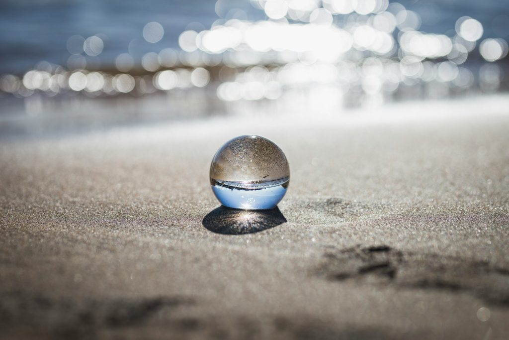 A glass ball lies on flat sand on beach, reflecting an upside-down blue sky. The glistening calm ocean gently laps the beach in the background.