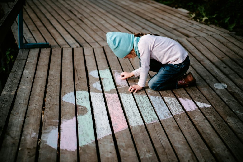 A child draws pink and blue hearts using chalk on a wooden boardwalk.