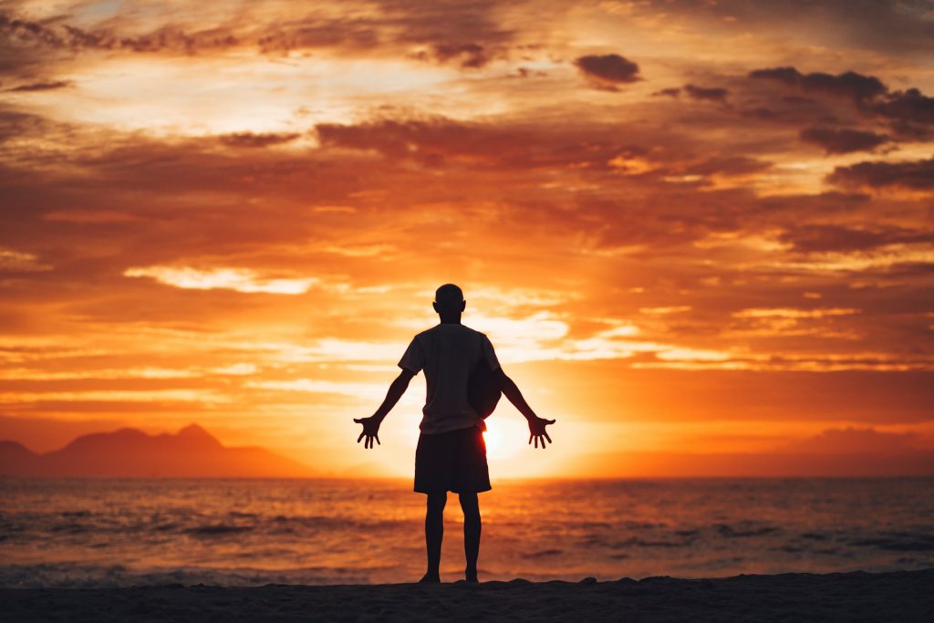 A man stands on a beach facing the water as the sun sets over the horizon.
