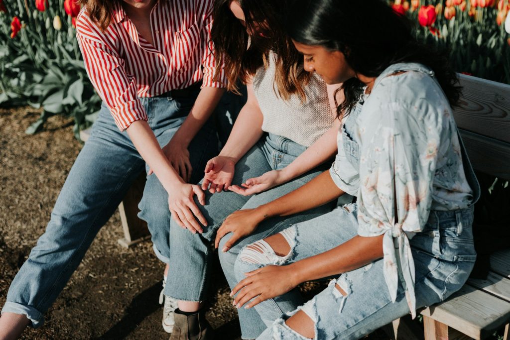 Three women sit on a park bench, two either side are laying hands on a third. All are in prayer.